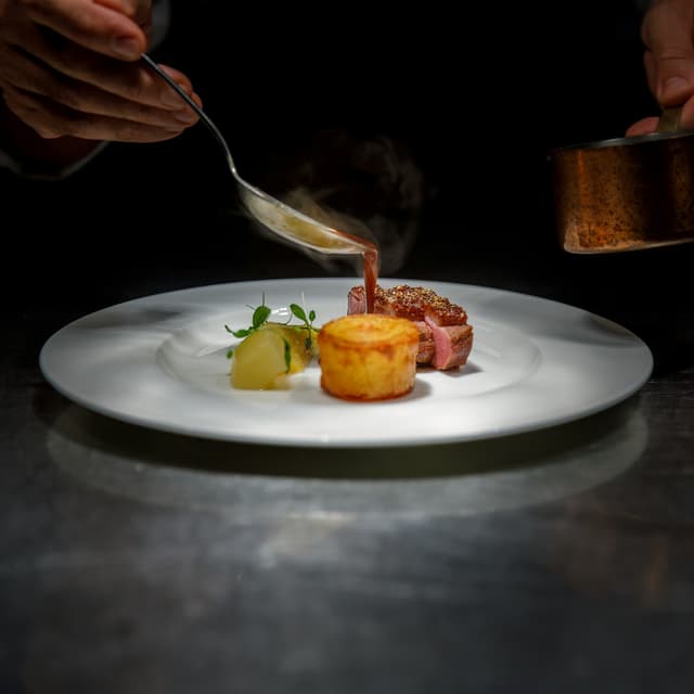 A chef plating a dish with sauce in a fine dining kitchen